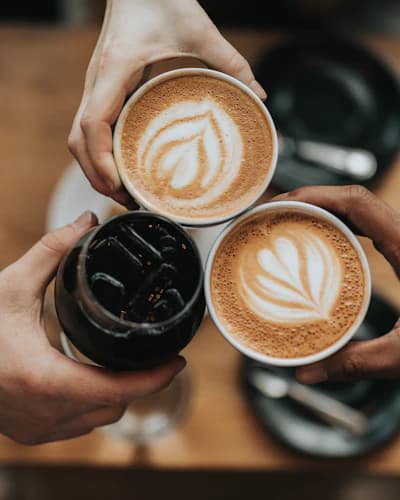 Três mãos brindando com copos de café sobre mesa de madeira